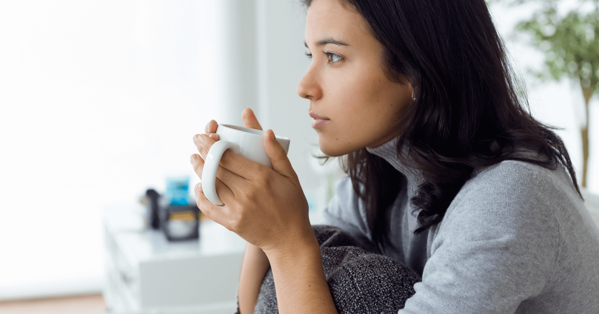 Woman holding a cup of coffee. Sitting with her knees pulled up to her chest and thinking as she looks into the distance.