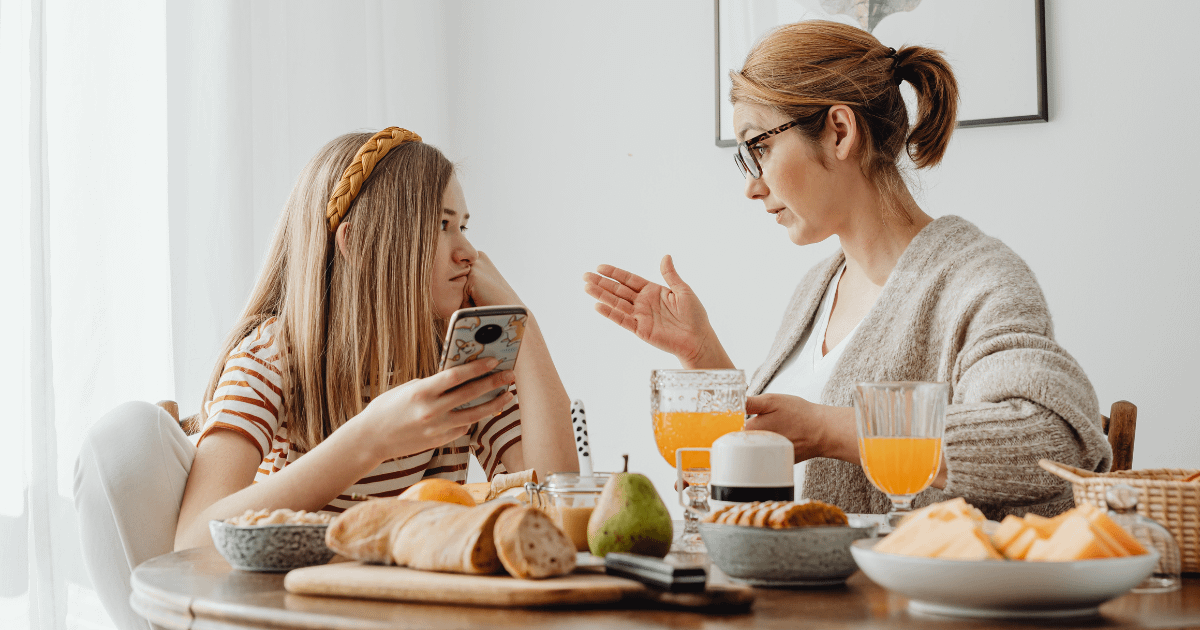 Mother and teen daughter are talking at the breakfast table. The teen girl has her cell phone in her hand and looks annoyed.