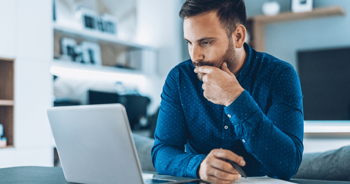 A man sits at his laptop at home, hand to his chin in thought, navigating the challenge of finding affordable mental health support for his child.