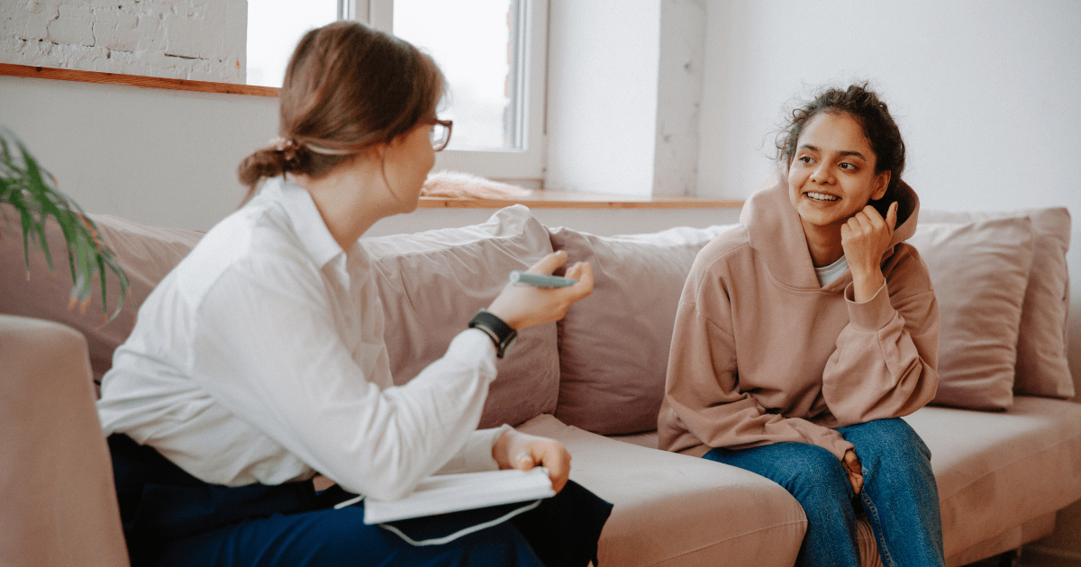 Photo of teen girl sitting on a couch talking with her therapist, who has a pen in her hand and a notebook in her lap.