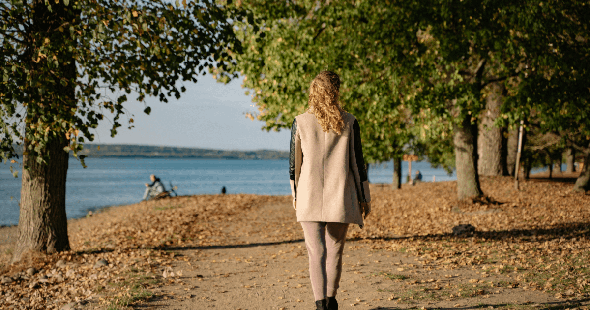 Woman walking alone on a tree-lined path near a lake, seen from behind, capturing the quiet isolation of parenting a struggling child.