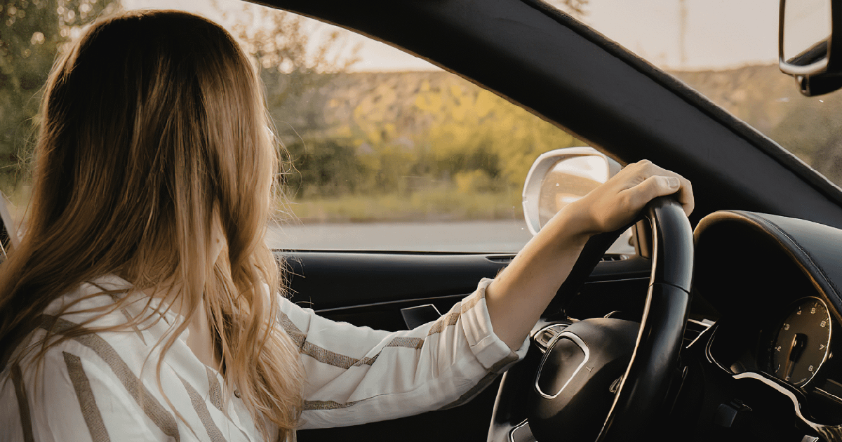 A woman driving alone, seen from behind, one hand on the steering wheel, with golden afternoon light coming through the windshield.