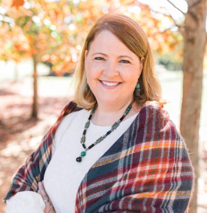 White woman standing amongst trees with her hands in front of her.